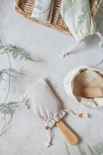A natural soap brush with a wooden handle is placed on a light textured surface, surrounded by a wicker basket and fabric pouches adorned with fern prints. One pouch appears to be open, revealing a powdery substance inside.