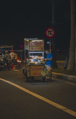 A street vendor dressed in a blue shirt and wearing a face mask stands beside a small food cart. The cart is illuminated and displays signs with Vietnamese text including 'Chả Cá Chiên Mắm' and 'Bắp Xào'. It's nighttime, and the scene is set on a dimly lit street with other people and distant lights visible in the background.