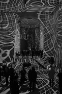 Intricate white grid-like patterns are projected onto the walls, floor, and ceiling of a large indoor space filled with people. The designs create a dynamic and surreal atmosphere, as they warp and wrap around the architectural features and people, adding depth and texture to the scene.