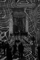 Intricate white grid-like patterns are projected onto the walls, floor, and ceiling of a large indoor space filled with people. The designs create a dynamic and surreal atmosphere, as they warp and wrap around the architectural features and people, adding depth and texture to the scene.