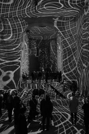 Intricate white grid-like patterns are projected onto the walls, floor, and ceiling of a large indoor space filled with people. The designs create a dynamic and surreal atmosphere, as they warp and wrap around the architectural features and people, adding depth and texture to the scene.