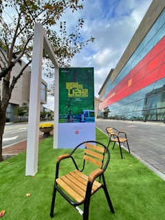 An outdoor setting featuring a poster stand displaying a green-themed poster with text in a foreign language. Wooden chairs with black metal frames are arranged on artificial green grass. A modern building with a glass facade and red accents is visible in the background, adjacent to a sidewalk and a street. Nearby, a small tree provides natural elements to the scene.
