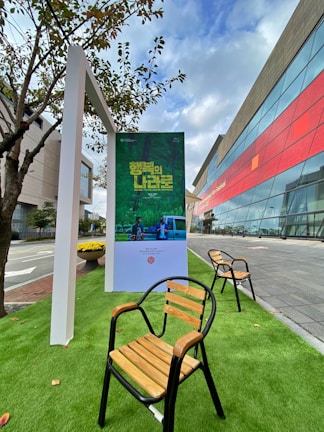 An outdoor setting featuring a poster stand displaying a green-themed poster with text in a foreign language. Wooden chairs with black metal frames are arranged on artificial green grass. A modern building with a glass facade and red accents is visible in the background, adjacent to a sidewalk and a street. Nearby, a small tree provides natural elements to the scene.