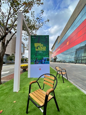 An outdoor setting featuring a poster stand displaying a green-themed poster with text in a foreign language. Wooden chairs with black metal frames are arranged on artificial green grass. A modern building with a glass facade and red accents is visible in the background, adjacent to a sidewalk and a street. Nearby, a small tree provides natural elements to the scene.