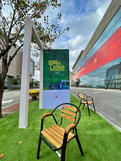 An outdoor setting featuring a poster stand displaying a green-themed poster with text in a foreign language. Wooden chairs with black metal frames are arranged on artificial green grass. A modern building with a glass facade and red accents is visible in the background, adjacent to a sidewalk and a street. Nearby, a small tree provides natural elements to the scene.