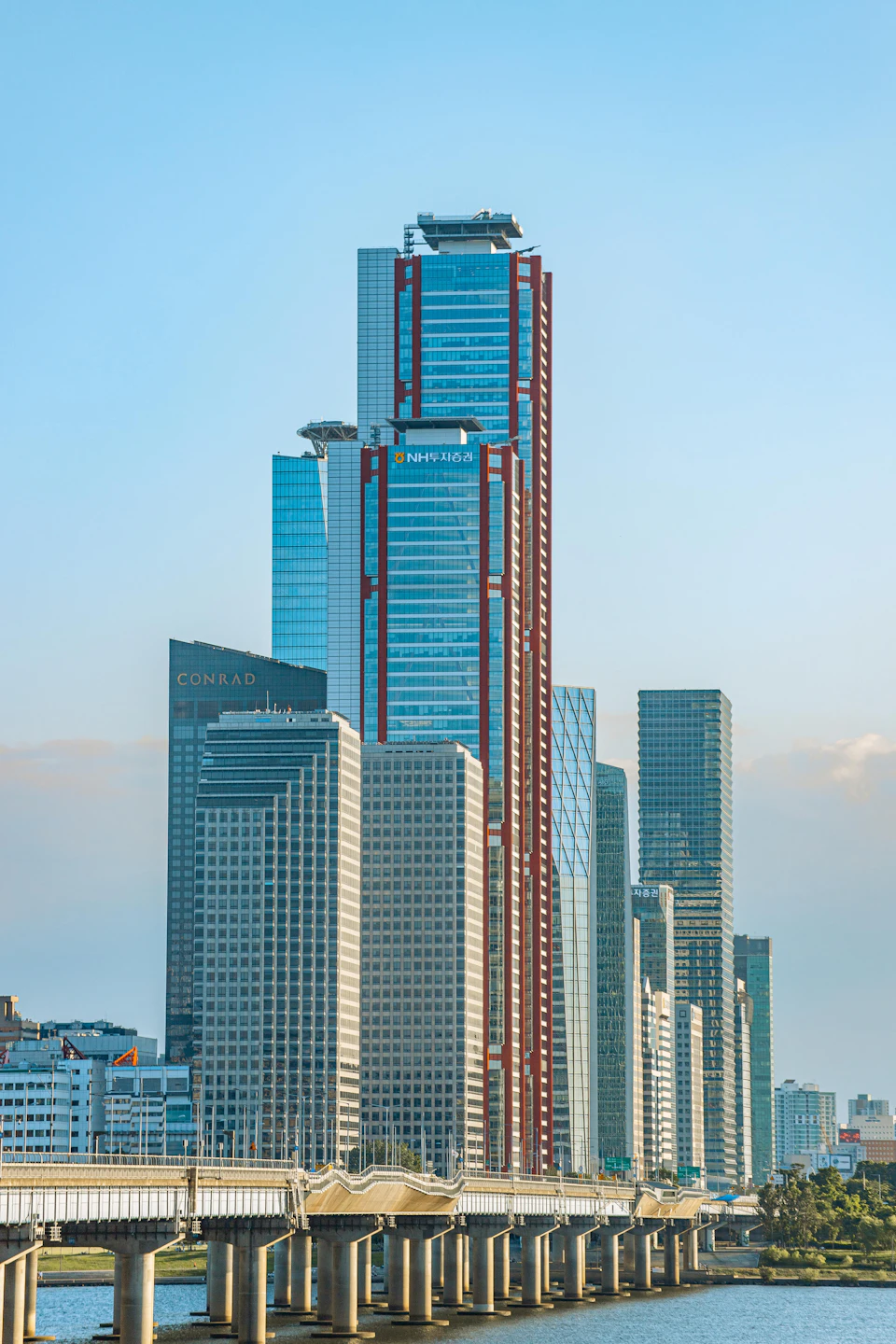 The image depicts a bustling cityscape with a prominent red and white skyscraper on the right side, towering over the surrounding buildings. The sky above is a clear blue, providing a beautiful backdrop to the urban scene below. A bridge can be seen in the foreground, connecting two parts of the city separated by water.