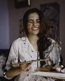 A person is smiling with joy, face and hands covered in various colors of paint. They are holding art supplies, including a palette and paint tubes. The background features a partially visible painting, suggesting an art studio setting.