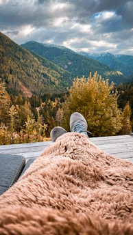 A serene mountain landscape on an overcast day, with lush green forests covering the hills. In the foreground, a person is relaxing on a wooden deck with their feet up, wrapped in a soft, fluffy brown blanket. The scene conveys a sense of tranquility and escape into nature.