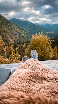 A serene mountain landscape on an overcast day, with lush green forests covering the hills. In the foreground, a person is relaxing on a wooden deck with their feet up, wrapped in a soft, fluffy brown blanket. The scene conveys a sense of tranquility and escape into nature.