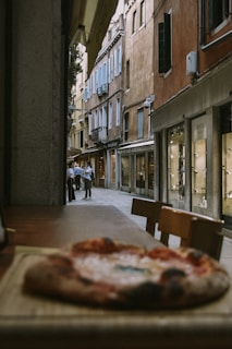 A cozy street scene with rustic buildings and blue shutters set in a narrow alleyway. A pizza sits on a wooden table in the foreground, while a group of people stand in conversation further down the street. The scene has a warm, inviting vibe with soft diffused lighting.
