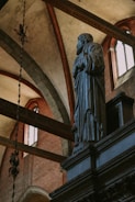 A stone statue of a robed figure with hands clasped is positioned inside a grand architectural space. The background features high vaulted ceilings with decorative arches, windows allowing in natural light, and intricate details. The atmosphere is serene and solemn.