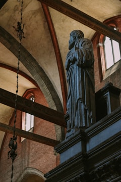 A stone statue of a robed figure with hands clasped is positioned inside a grand architectural space. The background features high vaulted ceilings with decorative arches, windows allowing in natural light, and intricate details. The atmosphere is serene and solemn.