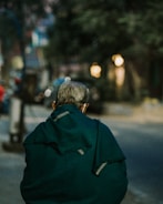 An elderly person is seen from behind walking along a street, wrapped in a dark green shawl. The surroundings are slightly out of focus, suggesting an urban setting with blurred lights indicating evening or early night time.