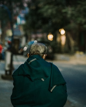 An elderly person is seen from behind walking along a street, wrapped in a dark green shawl. The surroundings are slightly out of focus, suggesting an urban setting with blurred lights indicating evening or early night time.