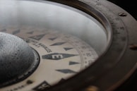 Close-up view of a vintage compass with a glass cover. The compass card displays cardinal points and degree markings, with visible text indicating 'N, E, S, W' directions. The outer rim has a metallic finish with detailed markings and screws.