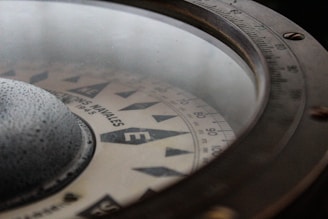 Close-up view of a vintage compass with a glass cover. The compass card displays cardinal points and degree markings, with visible text indicating 'N, E, S, W' directions. The outer rim has a metallic finish with detailed markings and screws.