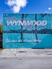 A mural painted on a building wall with the text 'Welcome to Wynwood Miami' adorned in large, stylized letters. The mural also features palm trees and the phrase 'Together We Create History' in a script font. The wall is predominantly teal, with elements of pink and purple in the text. The scene includes a glimpse of pavement and a small portion of sky with clouds.