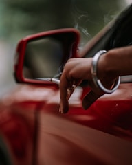 A close-up of a person's hand holding a cigarette out of a car window. The focus is on the hand and cigarette, with a red car door and side mirror visible. Smoke gently rises from the cigarette, adding a relaxed ambiance to the scene.