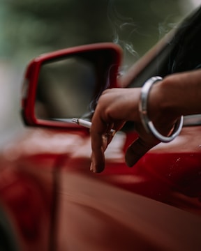A close-up of a person's hand holding a cigarette out of a car window. The focus is on the hand and cigarette, with a red car door and side mirror visible. Smoke gently rises from the cigarette, adding a relaxed ambiance to the scene.