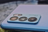 A close-up of a smartphone camera module with multiple lenses and text indicating '64MP AI Quad-Cam'. The phone features a sleek, metallic finish and is resting on a textured surface.