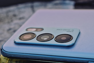 A close-up of a smartphone camera module with multiple lenses and text indicating '64MP AI Quad-Cam'. The phone features a sleek, metallic finish and is resting on a textured surface.