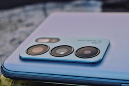 A close-up of a smartphone camera module with multiple lenses and text indicating '64MP AI Quad-Cam'. The phone features a sleek, metallic finish and is resting on a textured surface.