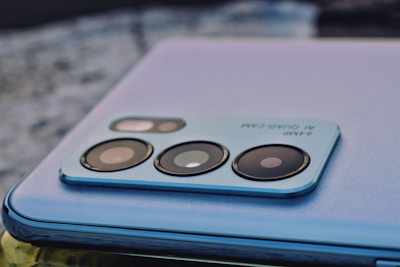 A close-up of a smartphone camera module with multiple lenses and text indicating '64MP AI Quad-Cam'. The phone features a sleek, metallic finish and is resting on a textured surface.