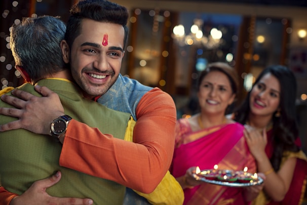A young man with a red tilak on his forehead hugs an older man wearing a wristwatch. Two women in colorful traditional clothing stand in the background, smiling and holding a decorated plate with lit candles. The setting appears festive with blurred lights in the backdrop.