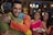 A young man with a red tilak on his forehead hugs an older man wearing a wristwatch. Two women in colorful traditional clothing stand in the background, smiling and holding a decorated plate with lit candles. The setting appears festive with blurred lights in the backdrop.