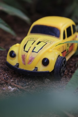 A toy car resembling a vintage Volkswagen Beetle. The car is primarily yellow with black accents and is situated on a dirt surface surrounded by green leaves, suggesting an outdoor setting.