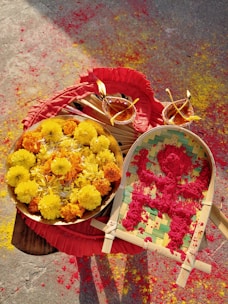 A beautifully arranged display of bright marigold flowers in yellow and orange hues sits on a wooden table. Next to it, a bamboo tray holds vibrant red powder. Decorative clay lamps with flame and colorful designs add a festive touch. Brightly colored powders are scattered on the ground, creating a lively and celebratory atmosphere.