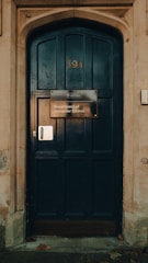 A large dark wooden door with an arched frame, featuring a sign that reads 'Department of Computer Science' from the University of Oxford. The door has the number 39A displayed above the sign and is surrounded by a stone wall. A few fallen leaves are scattered at the base.