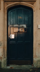 A large dark wooden door with an arched frame, featuring a sign that reads 'Department of Computer Science' from the University of Oxford. The door has the number 39A displayed above the sign and is surrounded by a stone wall. A few fallen leaves are scattered at the base.