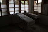 A dimly lit room contains a medical examination table covered with a white sheet. Wooden-framed windows line the wall, letting in diffused natural light. There are multiple bowls on a table to the left, and two woven baskets are placed on a table to the right, both draped with white cloth. The atmosphere is minimal and quiet.