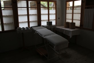 A dimly lit room contains a medical examination table covered with a white sheet. Wooden-framed windows line the wall, letting in diffused natural light. There are multiple bowls on a table to the left, and two woven baskets are placed on a table to the right, both draped with white cloth. The atmosphere is minimal and quiet.