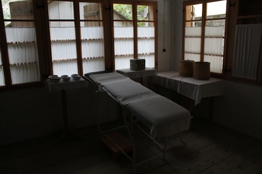 A dimly lit room contains a medical examination table covered with a white sheet. Wooden-framed windows line the wall, letting in diffused natural light. There are multiple bowls on a table to the left, and two woven baskets are placed on a table to the right, both draped with white cloth. The atmosphere is minimal and quiet.