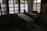 A dimly lit room contains a medical examination table covered with a white sheet. Wooden-framed windows line the wall, letting in diffused natural light. There are multiple bowls on a table to the left, and two woven baskets are placed on a table to the right, both draped with white cloth. The atmosphere is minimal and quiet.