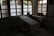 A dimly lit room contains a medical examination table covered with a white sheet. Wooden-framed windows line the wall, letting in diffused natural light. There are multiple bowls on a table to the left, and two woven baskets are placed on a table to the right, both draped with white cloth. The atmosphere is minimal and quiet.