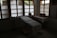 A dimly lit room contains a medical examination table covered with a white sheet. Wooden-framed windows line the wall, letting in diffused natural light. There are multiple bowls on a table to the left, and two woven baskets are placed on a table to the right, both draped with white cloth. The atmosphere is minimal and quiet.