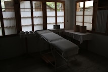 A dimly lit room contains a medical examination table covered with a white sheet. Wooden-framed windows line the wall, letting in diffused natural light. There are multiple bowls on a table to the left, and two woven baskets are placed on a table to the right, both draped with white cloth. The atmosphere is minimal and quiet.