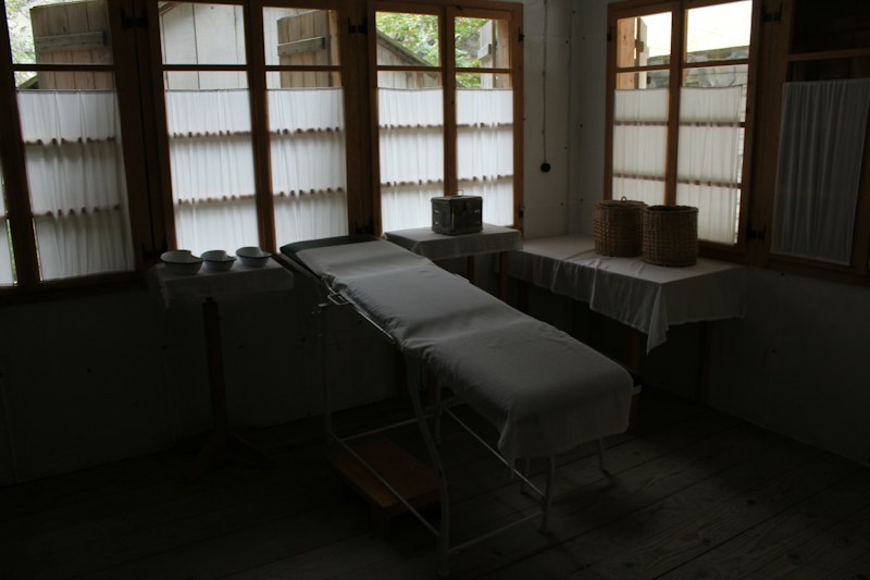 A dimly lit room contains a medical examination table covered with a white sheet. Wooden-framed windows line the wall, letting in diffused natural light. There are multiple bowls on a table to the left, and two woven baskets are placed on a table to the right, both draped with white cloth. The atmosphere is minimal and quiet.