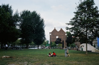 A park area with several people, including children playing on the grass, surrounded by trees. In the background, there is a distinctive brick building with a gabled roof, and some steps or ramps on the right side leading up to it.