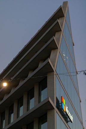 A modern architectural building with sharp angles and multiple glass windows reflecting the sky. The structure features a prominent logo of a well-known technology company on one side. The image is taken during dusk, as evident from the lighting and the darkening sky.
