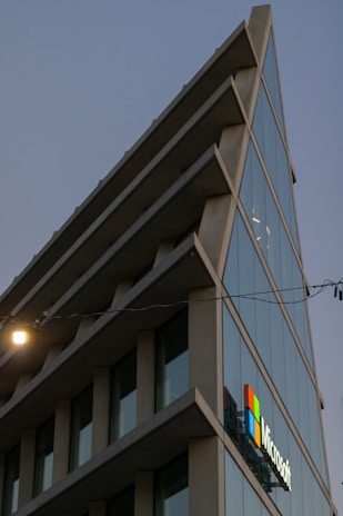 A modern architectural building with sharp angles and multiple glass windows reflecting the sky. The structure features a prominent logo of a well-known technology company on one side. The image is taken during dusk, as evident from the lighting and the darkening sky.