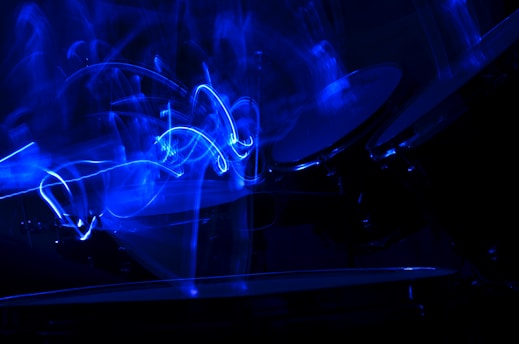 A visually striking image dominated by swirling blue light patterns, suggesting motion and energy. The abstract light trails create a dynamic composition against a dark background, with the faint outline of musical instruments like drums visible.