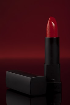 A vibrant red lipstick stands upright against a deep, red backdrop. The lipstick is partially extended from a sleek, black matte tube and is accompanied by a matching cap that lies horizontally beside it. The overall composition is elegant with a focus on the glossy texture of the lipstick.