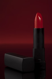 A vibrant red lipstick stands upright against a deep, red backdrop. The lipstick is partially extended from a sleek, black matte tube and is accompanied by a matching cap that lies horizontally beside it. The overall composition is elegant with a focus on the glossy texture of the lipstick.
