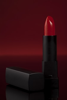 A vibrant red lipstick stands upright against a deep, red backdrop. The lipstick is partially extended from a sleek, black matte tube and is accompanied by a matching cap that lies horizontally beside it. The overall composition is elegant with a focus on the glossy texture of the lipstick.
