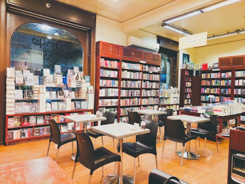 The scene shows a cozy bookstore with several black chairs and small tables arranged in the center of the room. Surrounding the seating area are tall wooden bookshelves filled with a variety of books. The shelves are neatly organized, and the room has a warm, inviting atmosphere. A large window is visible, allowing natural light to illuminate the space.