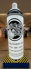 A large sculpture of an aerosol paint can with graffiti-style text on it is displayed in a shopping mall setting. The can features bold black and white colors with detailed graphic design elements. It sits on a platform with a yellow and black hazard pattern, indicative of industrial themes. The surrounding environment includes mall storefronts and overhead lighting.